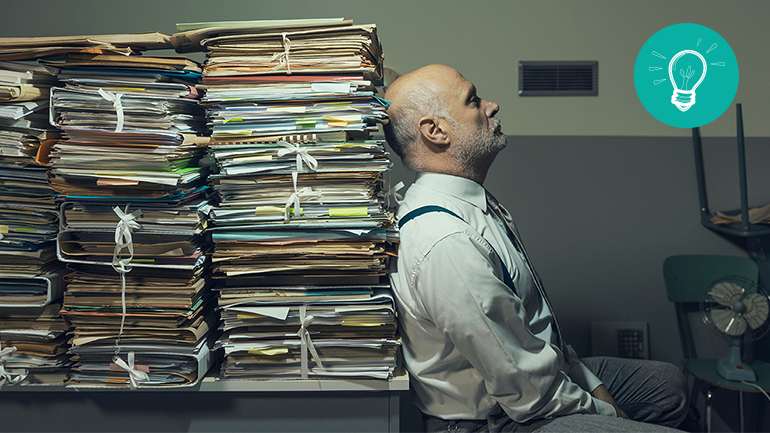 Mappar med papper i travar och en man som lutar sig mot dem och ser uppgiven ut.