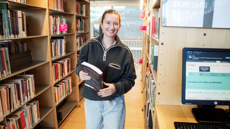 En person står i ett bibliotek och håller i en bok.