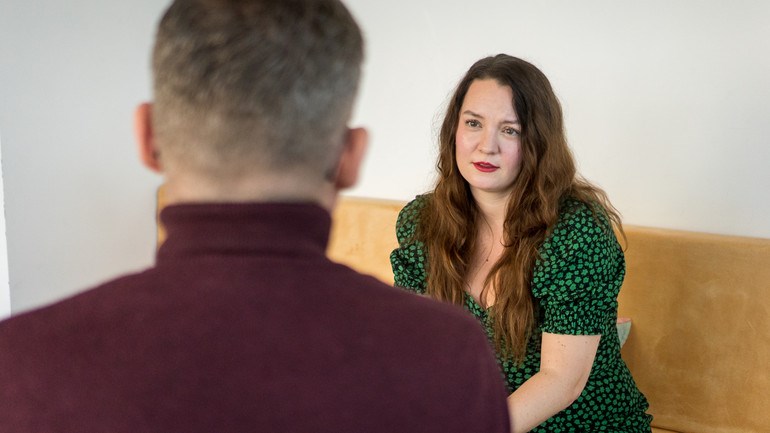En person sitter på en bänk och pratar med en annan person.