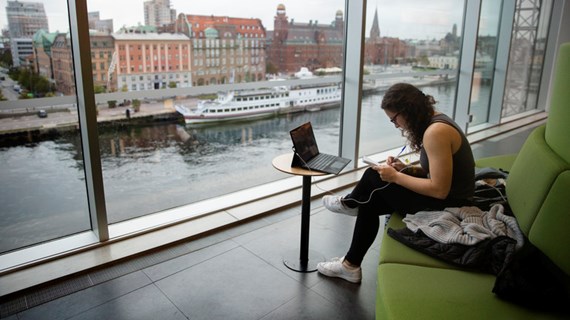 Student med anteckningsblock i handen och hörlurar i öronen pluggar framför stort fönster i Orkanenbiblioteket. 